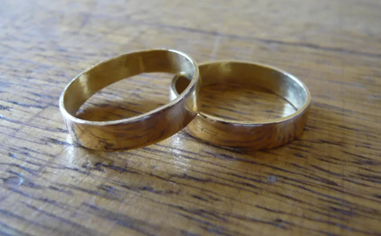 Primary relationship: two plain gold wedding rings resting together on a warm wooden table, a quiet still life about a marriage whose shape has changed